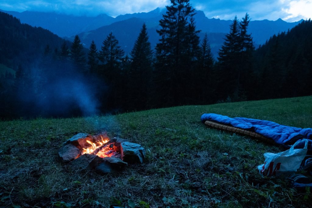 overlooking trees into the mountains, a sleeping bag on the ground and a fire burning