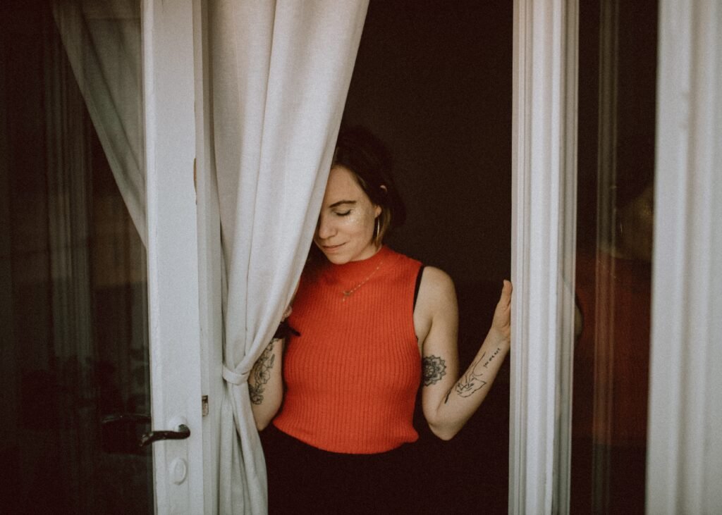 white woman in a red sleeveless sweater standing in  a doorway with white curtains. Her eyes are closed and she has tattoos on her arms.