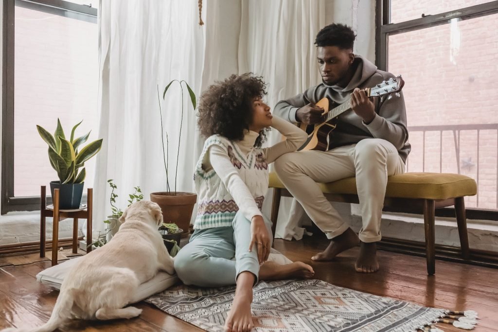 Full body of black boyfriend playing acoustic guitar for African American girlfriend while spending time together in room with dog