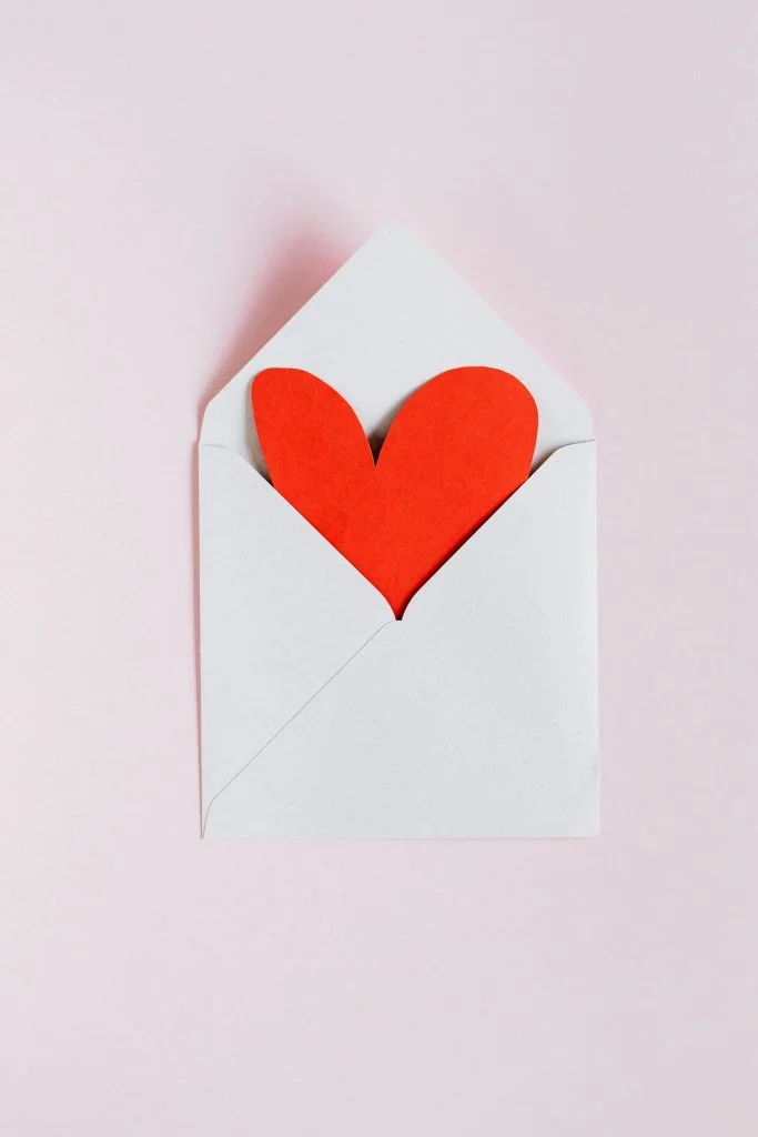 Top view of opened white envelope with handmade red paper heart placed against pink background as a scavenger hunt date night clue