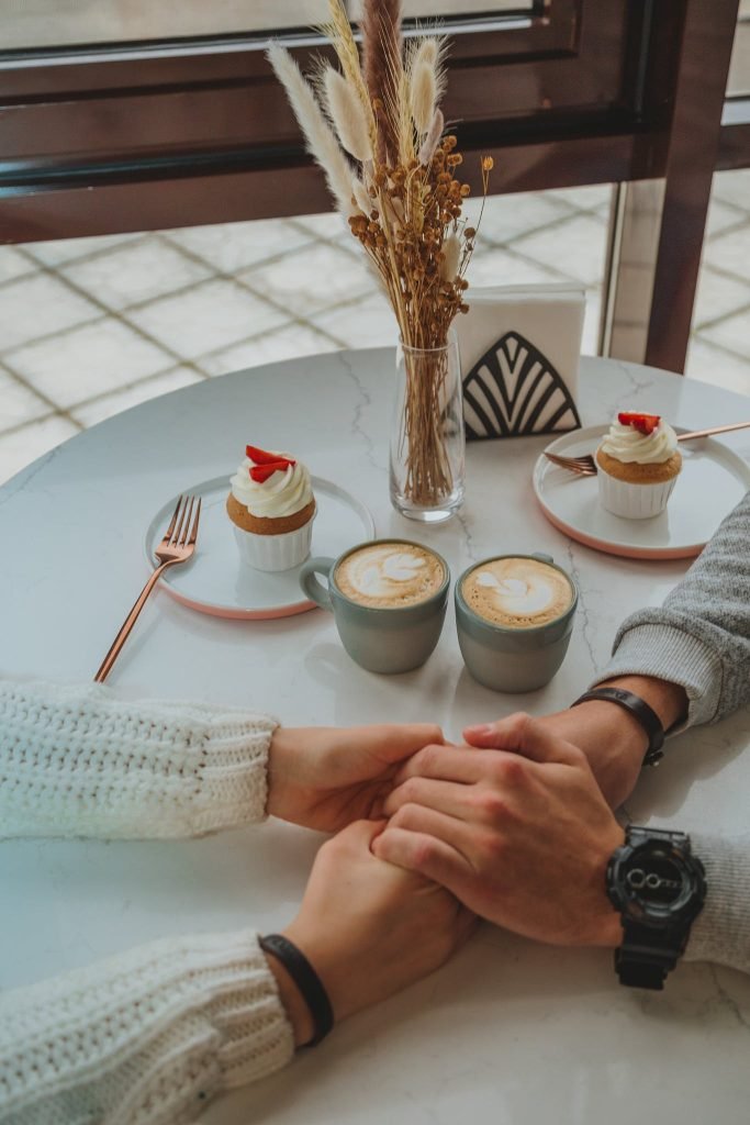 From above of unrecognizable romantic couple sitting near window at table with cups of latte and tasty cupcakes during date in restaurant