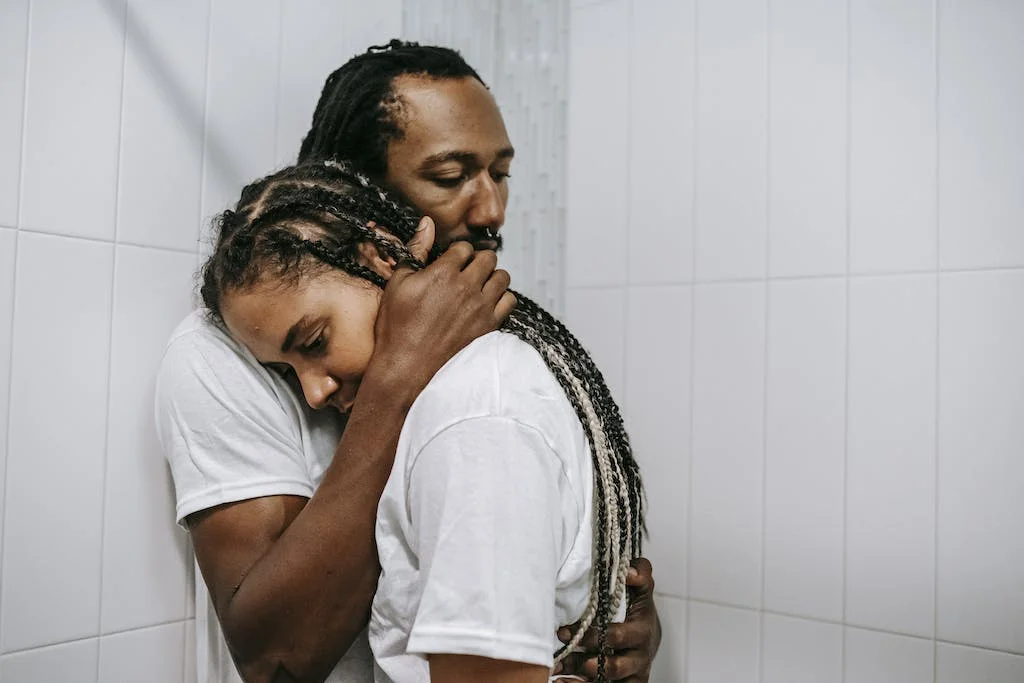 Stressed sad African American couple in white shirts embracing each other in light bathroom