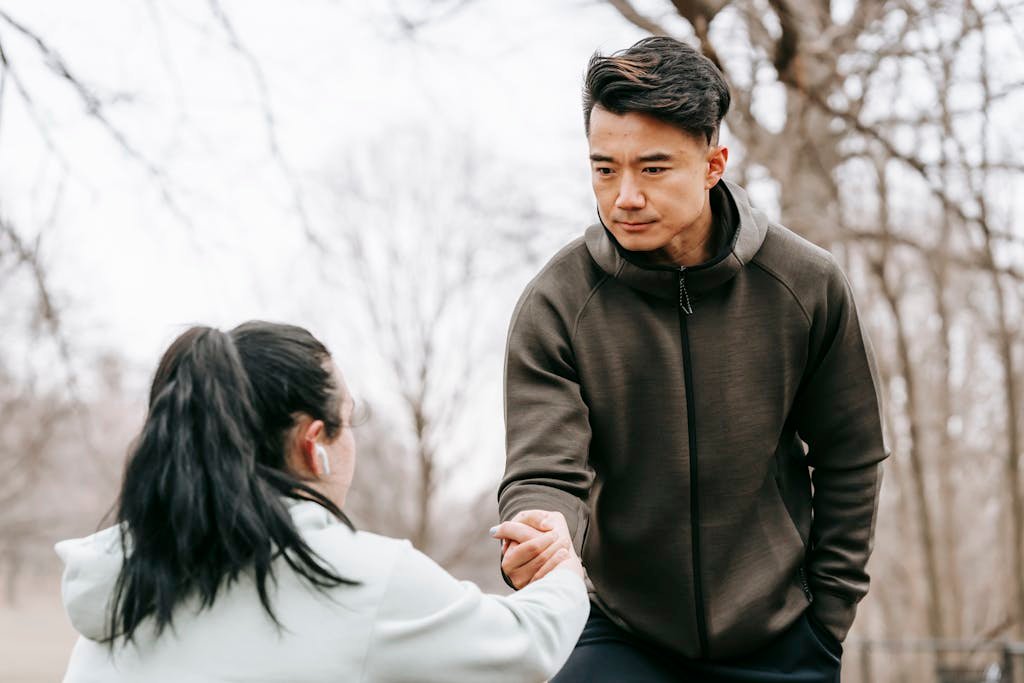 Calm Asian man in sportswear giving hand to unrecognizable female with dark hair in earphones in autumn park in daytime