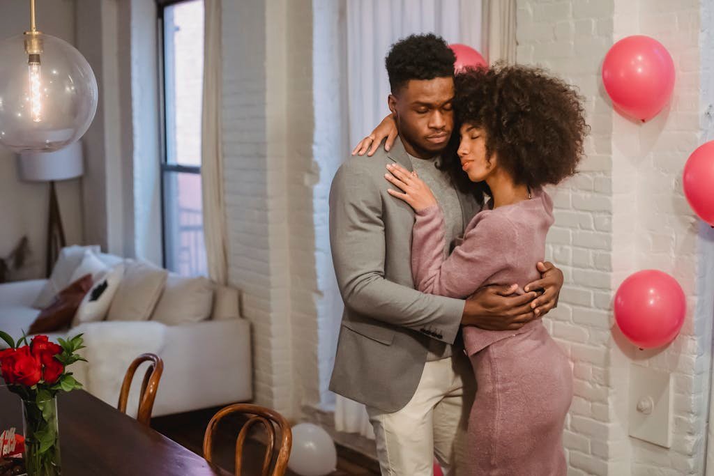 Dreamy young black couple in elegant clothes standing in light room and hugging while dancing together at brick wall with pink balloons