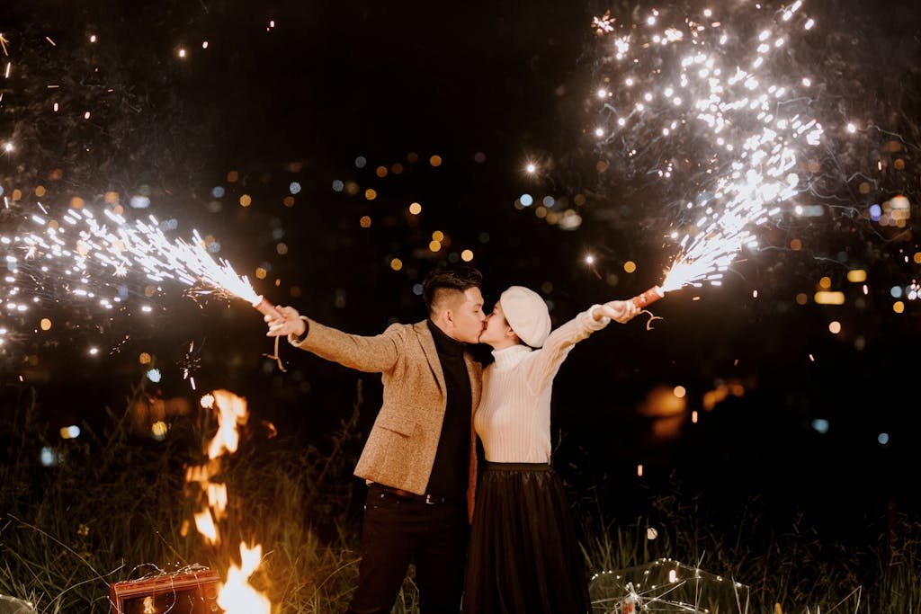 Happy ethnic couple kissing each other while holding burning fireworks near fire at night