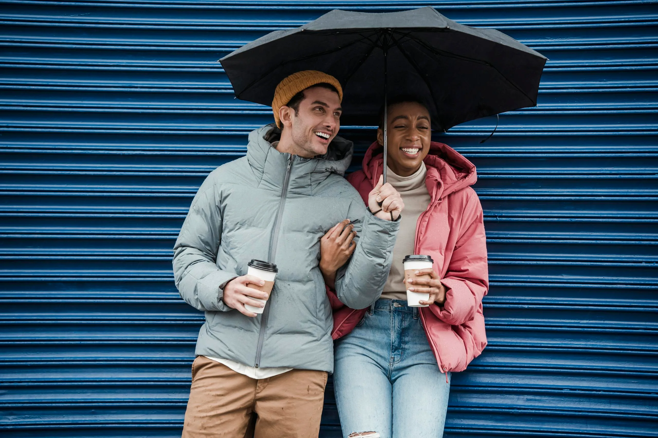 Happy multiracial couple with umbrella in warm outerwear laughing while standing on street with takeaway beverages in cold rainy weather