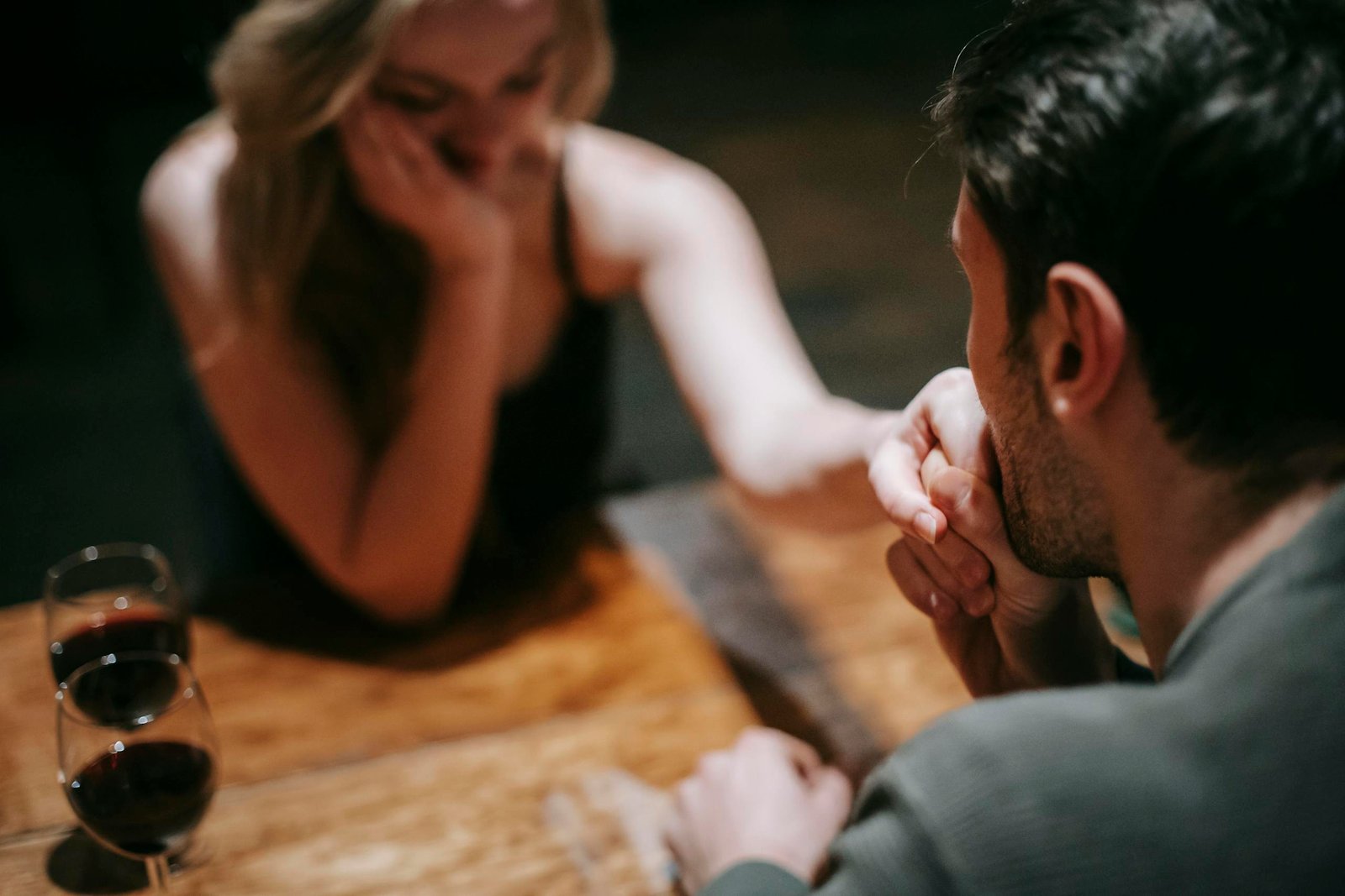 Anonymous young couple in elegant clothes sitting at wooden table with glasses with red wine while man kissing hand to beloved in light restaurant on anniversary date ideas