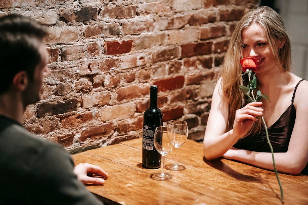 Young couple in elegant clothes sitting at wooden table near brick wall in light restaurant while enjoying date with red rose and wine near glasses and looking at each other