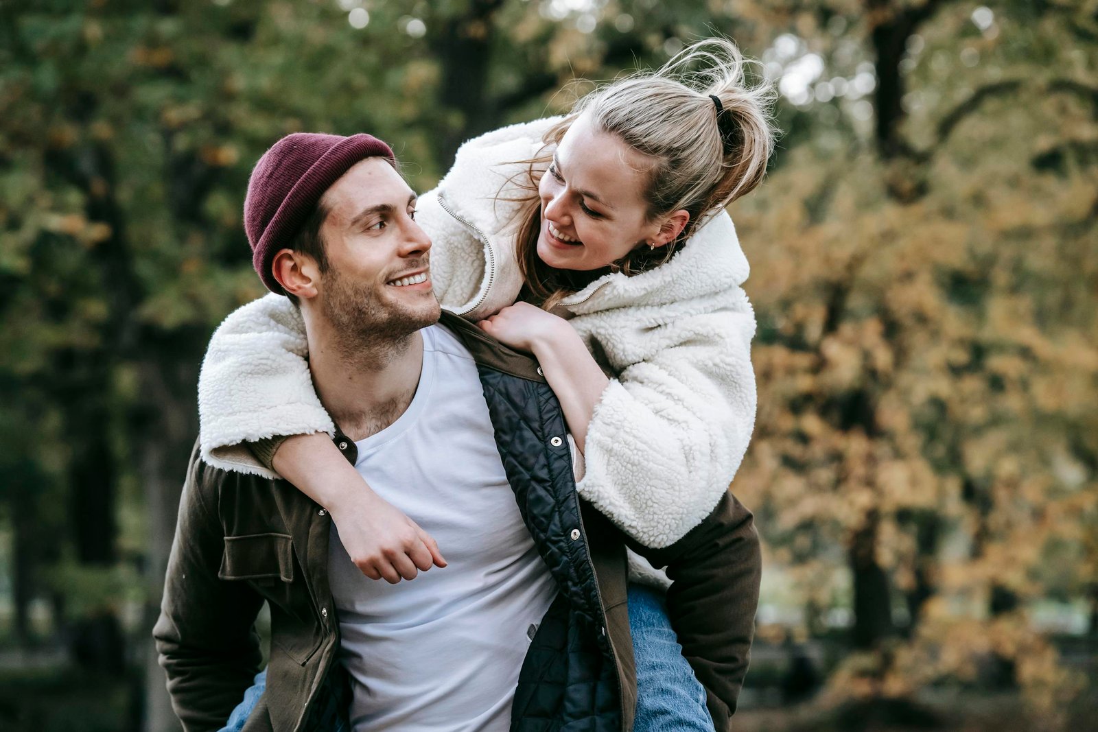 Young satisfied female on back of positive man in casual clothes spending time in autumn park