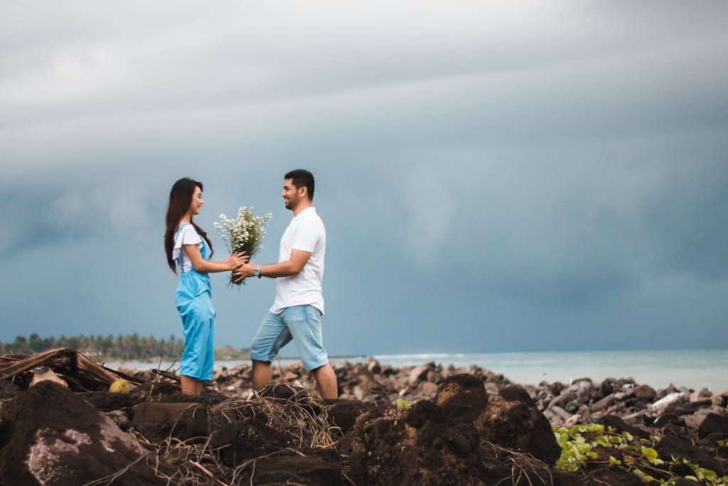 A couple enjoys a romantic moment by the rocky seashore under a cloudy sky.