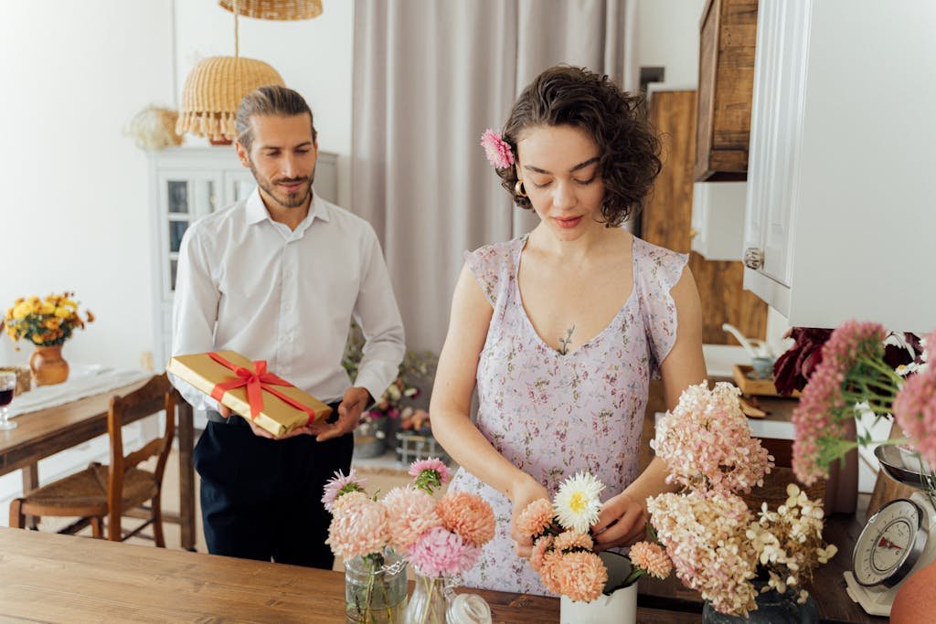 A couple shares a romantic moment as the man offers a gift while the woman arranges flowers in a cozy kitchen.