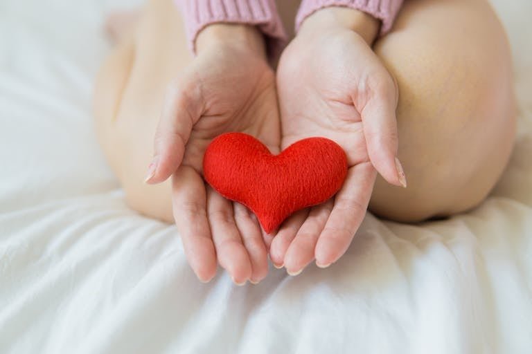 Unrecognizable female sitting with bare legs on white sheet with small red heart in hands in light room in daytime benefits of empathy