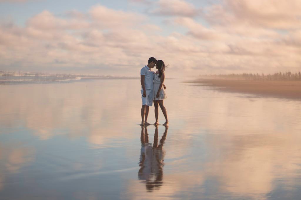 A couple embraces on a serene beach during sunset, reflecting on wet sand.