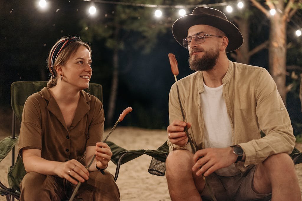 A couple enjoying a campfire at night, sitting in folding chairs and roasting food under string lights.