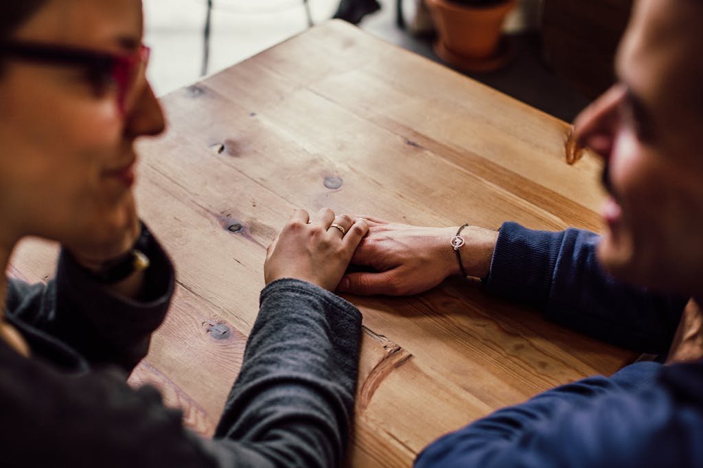 A couple shares a romantic moment holding hands across a wooden table indoors.