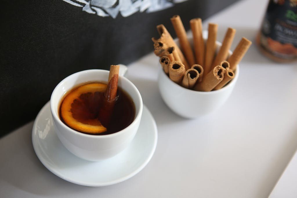 A white cup of cinnamon-flavored tea with a citrus slice, accompanied by cinnamon sticks.