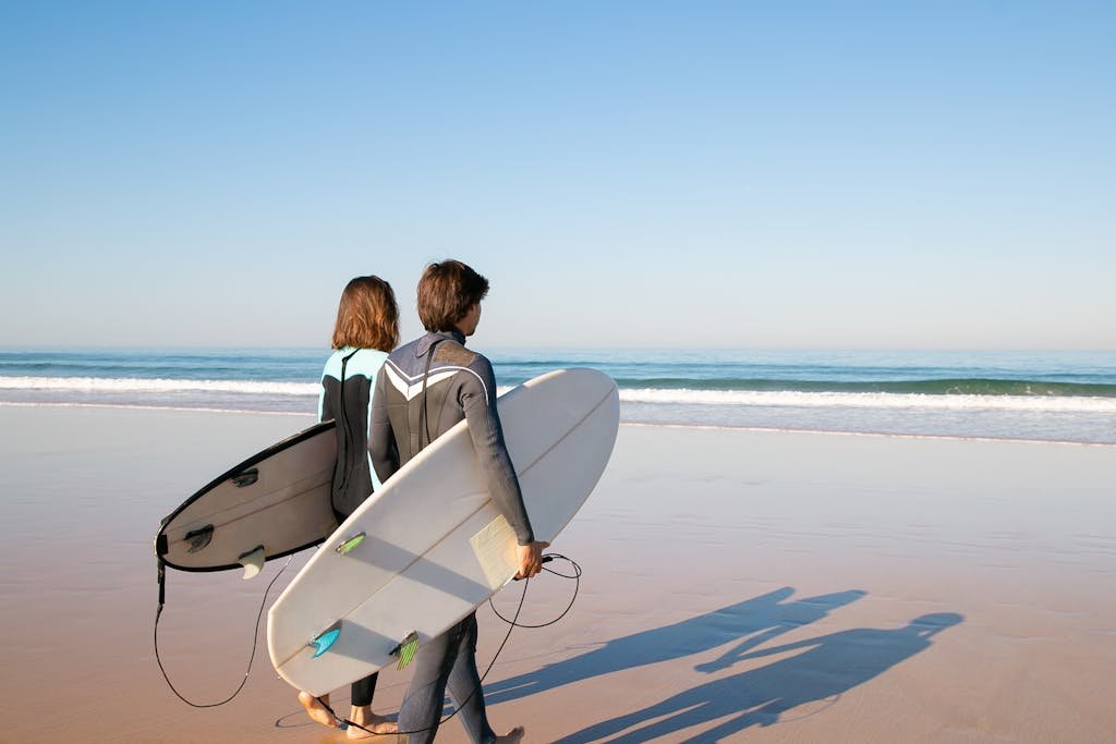 A young couple in wetsuits carrying surfboards along a sunny beach in Portugal.