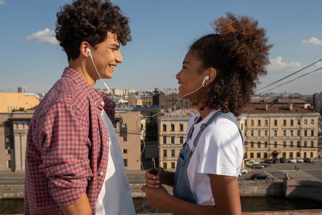 Two teenagers enjoy music together with shared earphones on a sunny rooftop.