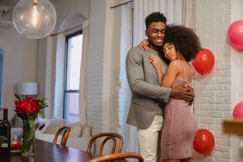 A couple embraces lovingly in a home setting adorned with roses and balloons.