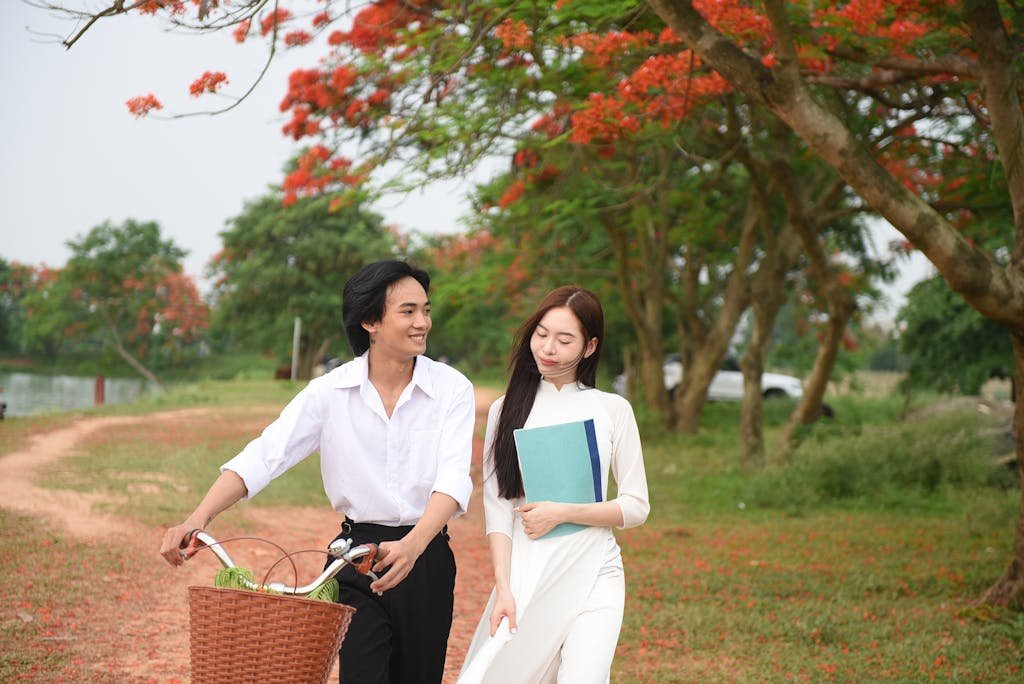 A couple enjoys a charming walk with a bicycle under vibrant red flowering trees in a park.