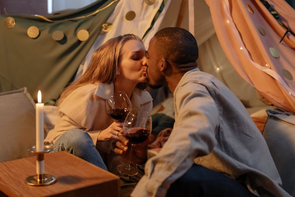 A couple shares a romantic kiss while holding wine glasses in cozy indoor setting.