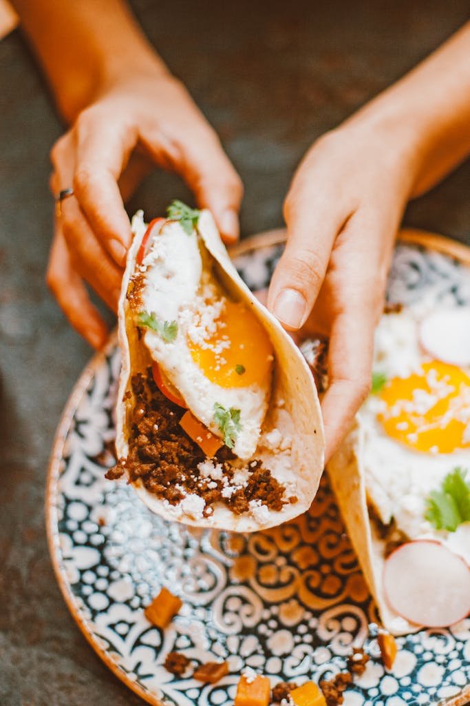 Close-up of a hand holding a breakfast taco with egg, beef, vegetables, and a colorful ceramic plate.