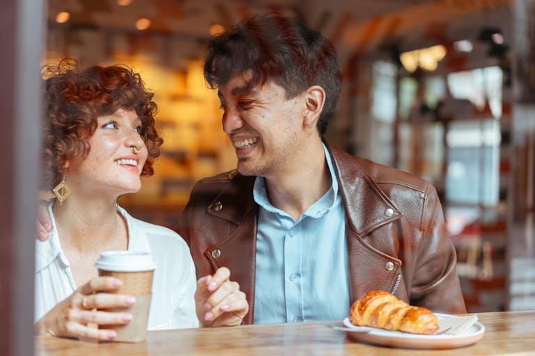 Couple laughing and enjoying coffee and croissant at a cozy cafe.