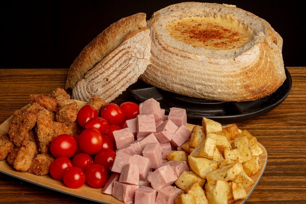 Delicious rustic bread bowl filled with cheese, surrounded by deli meats and fresh vegetables.