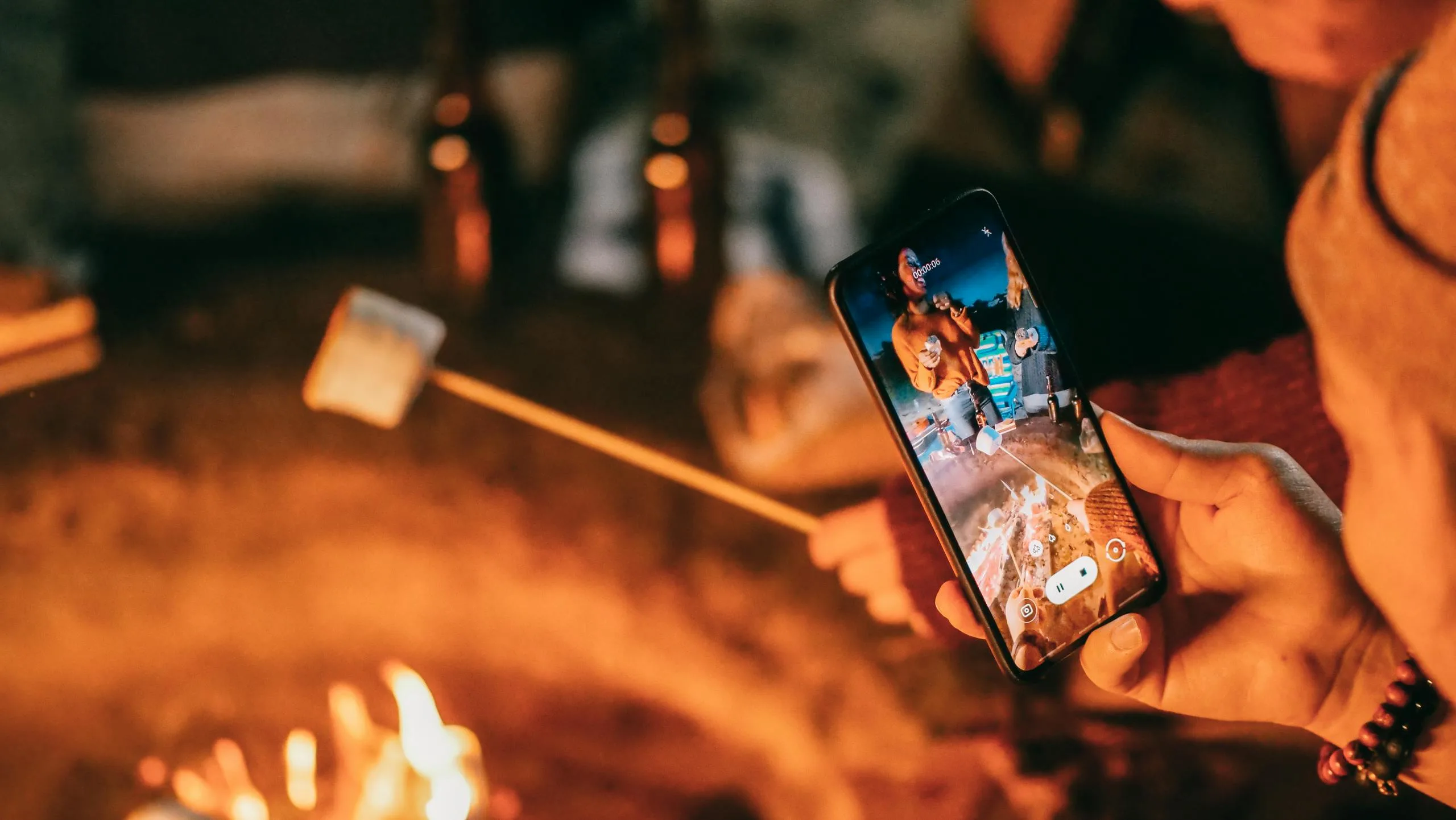 Friends enjoying a cozy campfire, roasting marshmallows and capturing moments on a smartphone.