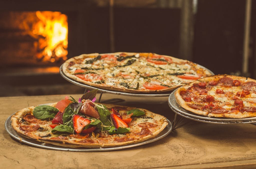 Trio of gourmet pizzas with unique toppings near a wood-fired oven, ideal for food enthusiasts.