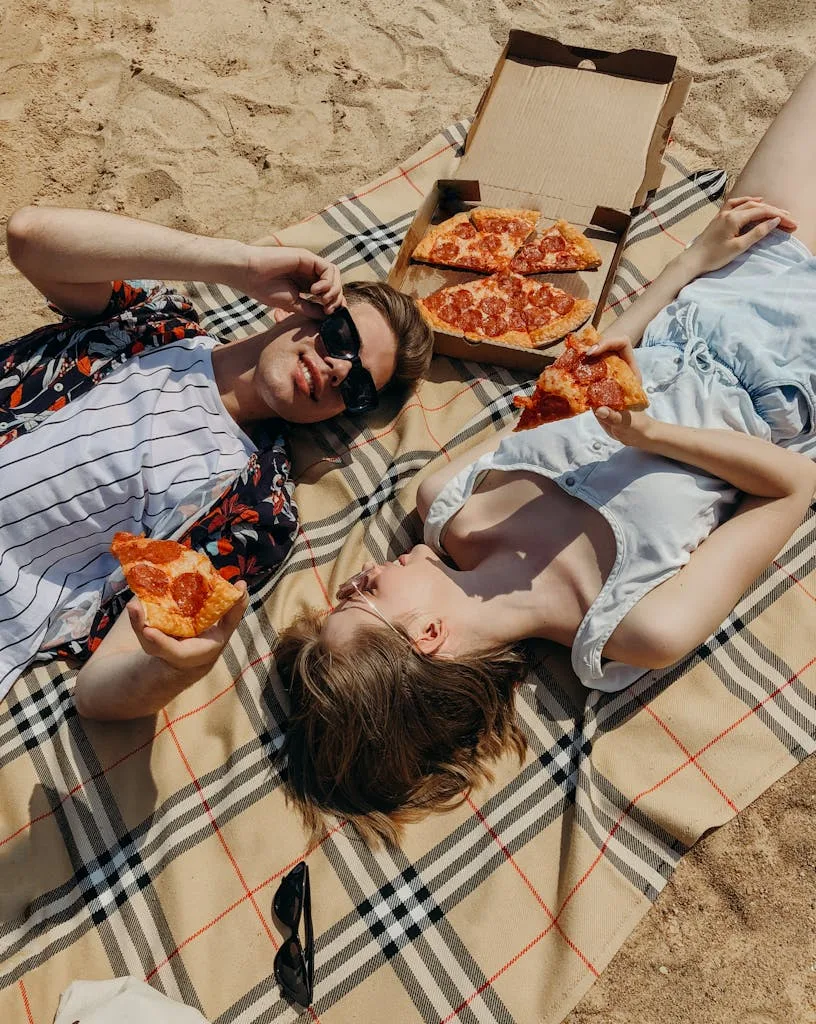 A young couple enjoying a sunny day at the beach with a pizza picnic.