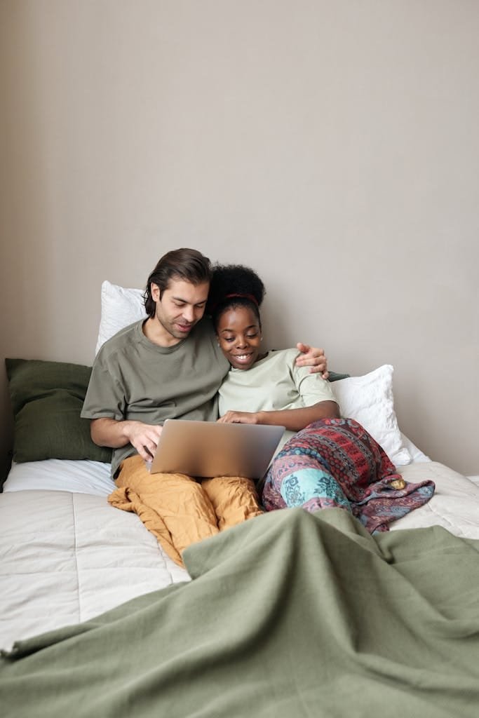 Couple enjoying leisure time, embracing while using a laptop on a comfortable bed.