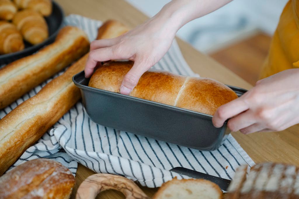 Hands holding a freshly baked loaf of bread in a pan, surrounded by various breads.