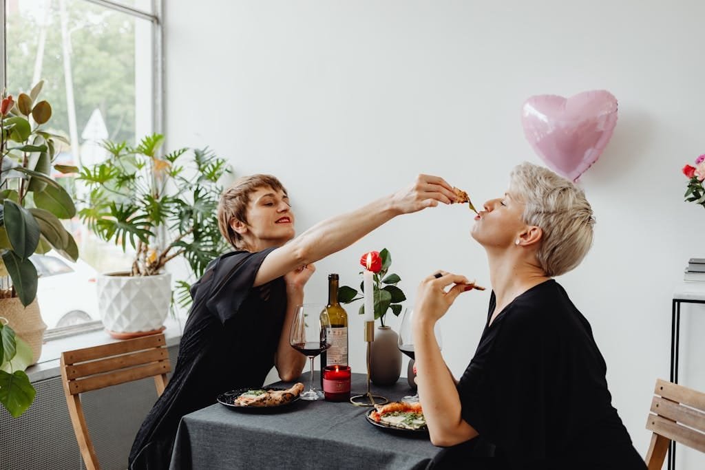A loving lesbian couple enjoying a romantic meal indoors with wine and a heart-shaped balloon.