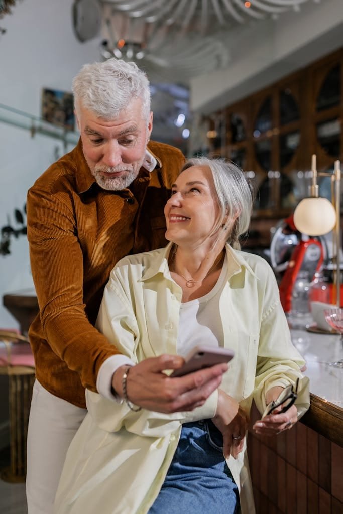 Happy senior couple using a smartphone together at a cozy indoor setting, sharing a joyful moment.